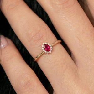 May include: A gold ring featuring a central oval-cut ruby surrounded by a halo of small, round diamonds. The ring's band is slender and polished, creating a delicate and elegant appearance. The ring is shown on a finger.