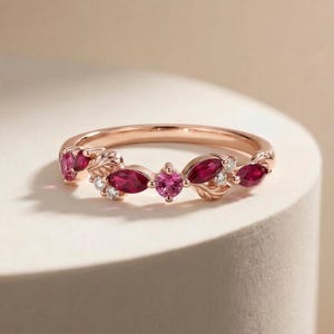 May include: Rose gold ring with a leaf design, featuring oval and round red gemstones and small white accent stones. The ring is set on a white surface with a soft background.