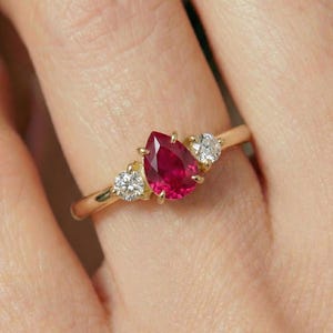 May include: A gold ring featuring a pear-shaped, vibrant red ruby center stone, flanked by two smaller, round, clear diamonds. The ring's band is a simple, polished gold. The ring is shown on a finger.