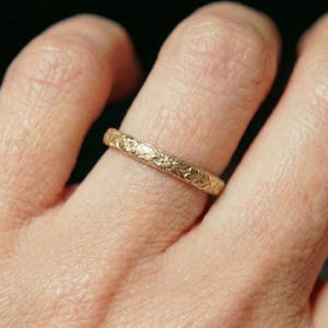 May include: A gold-colored ring with a floral pattern is shown on a finger. The ring has a continuous design of small flowers and leaves around its band. The ring is made of a shiny, yellow metal.