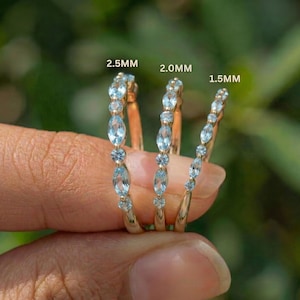 Peut inclure: Trois bagues en or serties de pierres précieuses bleu clair sont tenues dans une main. Les bagues sont étiquetées avec les tailles 2,5 mm, 2,0 mm et 1,5 mm. Les pierres précieuses sont ovales et rondes, serties dans une monture en or.