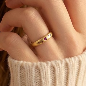 May include: A gold ring with small, embedded red gemstones and tiny star-like details. The ring is worn on a finger, with a cream-colored sweater in the background. The ring is a piece of jewelry.