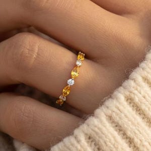 Puede incluir: Un anillo de oro con piedras preciosas amarillas y blancas alternas. El anillo está engastado con piedras amarillas de talla ovalada y piedras blancas redondas. El anillo se lleva en un dedo.