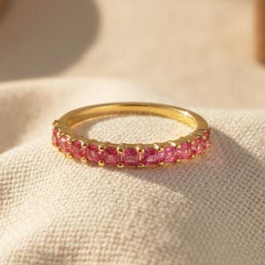 Könnte beinhalten: Ein goldener Ring mit einem Band aus kleinen, quadratisch geschliffenen rosa Edelsteinen. Der Ring ist vor einem neutralen Stoffhintergrund mit weicher Beleuchtung und unscharfem Hintergrund platziert.