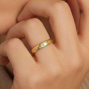 May include: Gold-toned ring with a series of small, round, turquoise-colored accents. The ring is a simple band design, with the turquoise accents evenly spaced around the circumference. The ring is worn on a finger.