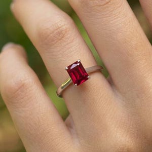 May include: A silver ring featuring a rectangular, deep red gemstone. The ring is set on a hand, showcasing the vibrant color of the stone. The design is simple and elegant, suitable for various occasions.