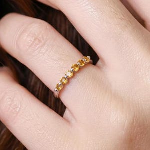 Peut inclure: Une bague en métal argenté ornée d'une rangée de sept petites pierres précieuses rondes de couleur jaune orangé, fixées par des griffes. La bague est portée au doigt, mettant en valeur l'anneau métallique et les pierres circulaires alignées.