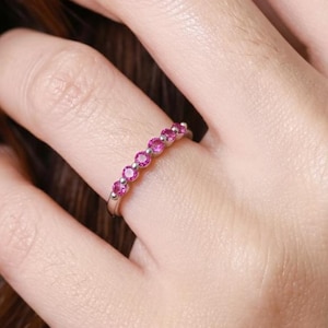 May include: A silver-toned band ring featuring a row of six small, round, vibrant pink gemstones set in a prong setting. The ring is worn on a finger, showcasing the circular cut of the stones and the polished metal finish of the band.