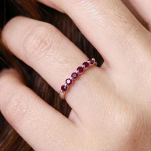 May include: A silver ring with seven round, vibrant red gemstones set in a row. The ring is displayed on a finger, showcasing the gemstones' color and the ring's delicate design. The ring is a piece of jewelry.