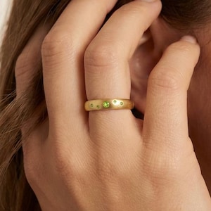 May include: A gold ring with a matte finish, adorned with small, round, green gemstones. The ring is worn on a finger, with the hand and part of the person's face visible in the image. The ring's design is simple yet elegant.