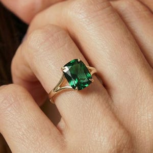 May include: A gold ring featuring a large, emerald-cut green gemstone. The ring has a delicate, split-shank design. The ring is shown on a person's finger.