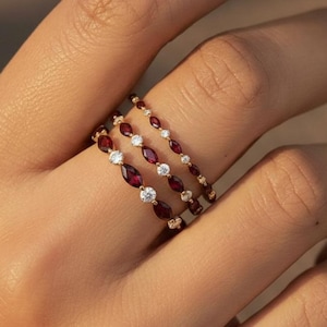 Op de afbeelding: Vier gouden ringen met edelstenen. Drie ringen hebben afwisselend rode en heldere edelstenen, terwijl de vierde ring heldere edelstenen heeft. De ringen zijn op een vinger gestapeld.