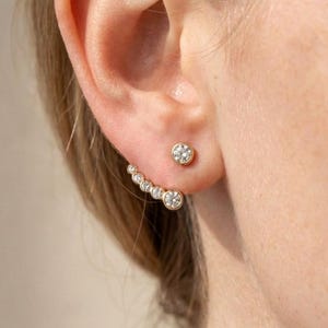 May include: Gold-toned earrings with clear, round gemstones. One earring is a single stud, while the other features a curved design with multiple stones. The earrings are displayed on an ear, showcasing their design and sparkle.