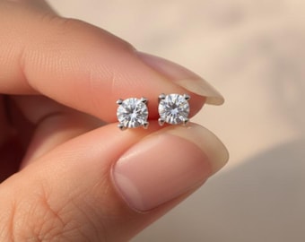 Puede incluir: Un par de pendientes de botón redondos y transparentes, con talla brillante y montados en un engaste de metal plateado de cuatro garras. Los pendientes presentan un diseño facetado que refleja la luz, creando un aspecto brillante.