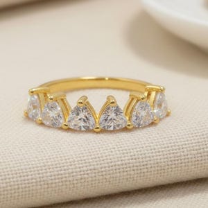 Puede incluir: Un anillo dorado con piedras preciosas transparentes talladas en forma de triángulo. El anillo está sobre una tela beige, con un plato blanco y un objeto dorado en el fondo. El diseño del anillo es elegante y llamativo.