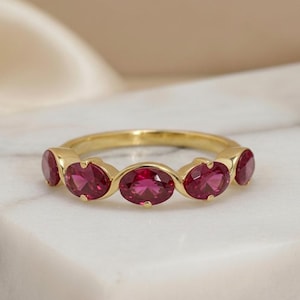 May include: A gold ring featuring a series of oval-cut, vibrant red gemstones. The gemstones are set in a continuous row along the ring's band. The ring is displayed on a white marble surface, with a soft, neutral background.