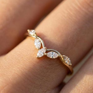 Könnte beinhalten: Ein goldfarbener Ring mit geschwungenem Design, besetzt mit mehreren klaren Edelsteinen im Marquise-Schliff. Der Ring wird an einem Finger getragen und zeigt die filigranen Details des Schmucks.