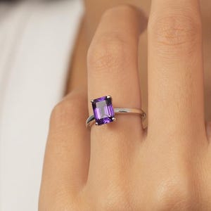 Op de afbeelding: Een zilveren ring met een rechthoekige, dieppaarse edelsteen. De ring is gezet op een slanke band, die de levendige kleur van de steen benadrukt. De ring wordt op een vinger getoond.