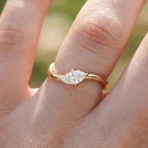 May include: A gold ring with a marquise-cut diamond. The ring has a twisted band design and a polished finish. The diamond is set horizontally in the center of the ring. The ring is shown on a finger.