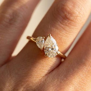 May include: A gold ring featuring two pear-shaped diamonds. The larger diamond is positioned centrally, with a smaller diamond adjacent to it. The ring's band is a thin, polished gold. The diamonds sparkle, reflecting light.