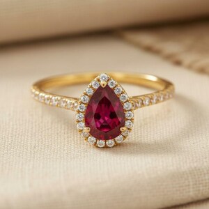 Puede incluir: Anillo de oro con una gema roja en forma de pera, rodeada por un halo de pequeños diamantes. La banda del anillo también está adornada con pequeños diamantes. El anillo se encuentra sobre un fondo neutro y texturizado.