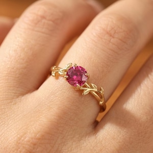 Puede incluir: Un anillo de oro con una gema rosa brillante. El anillo tiene una banda decorada con diseños de hojas. La gema es el elemento central, sujeta por garras, creando una pieza de joyería inspirada en la naturaleza.