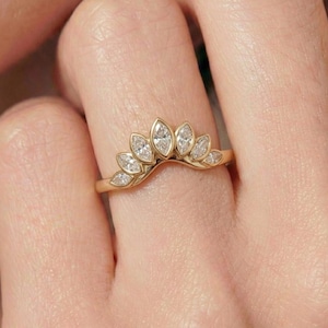 Puede incluir: Un anillo de oro con un diseño curvo, con siete diamantes talla marquesa. Los diamantes están engastados en un delicado patrón en forma de hoja, creando una apariencia de corona. El anillo se muestra en un dedo.