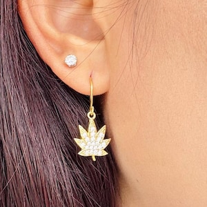 Op de afbeelding: Goudkleurige oorhangers met een cannabisbladontwerp, bezet met heldere kristallen. De oorbellen hangen aan een kleine gouden haak. Een kleine, ronde, heldere oorbel is ook zichtbaar in het oor.
