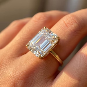May include: A gold ring featuring a large, rectangular emerald-cut clear gemstone held in place by four prongs. The band is a simple, smooth gold metal, and the stone reflects light with clear, linear facets across its surface.