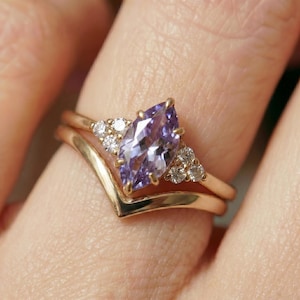 Puede incluir: Un conjunto de anillos de oro con una piedra preciosa morada clara con corte marquesa, flanqueada por piedras preciosas blancas más pequeñas. El conjunto incluye una banda en forma de cheurón. El anillo se muestra en un dedo.