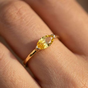 May include: A gold ring featuring a marquise-cut yellow gemstone. The gemstone is set in a delicate gold band, creating a simple yet elegant design. The ring is shown on a person's finger, highlighting its size and shape.