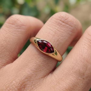Puede incluir: Un anillo de oro con una piedra preciosa roja oscura con corte marquesa. La piedra preciosa está engastada en un bisel y la banda es de oro liso y pulido. El anillo se muestra en un dedo, con un fondo verde borroso.
