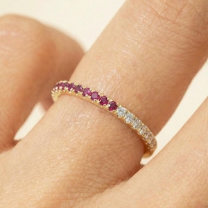 May include: A gold band ring set with alternating red and white gemstones. The ring is on a finger, showcasing the delicate design and vibrant colors of the gemstones. The ring is a piece of jewelry.