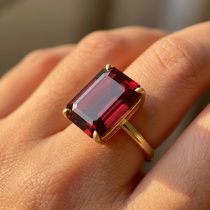 May include: A gold ring featuring a large, rectangular, deep red gemstone. The gemstone is set in a gold band with a simple, elegant design. The ring is shown on a finger.
