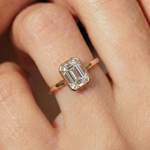 May include: A gold ring featuring a rectangular emerald-cut clear gemstone set in a smooth bezel setting. The band is thin and polished, showcasing a clean, minimalist design that highlights the faceted stone and the warm metallic finish.