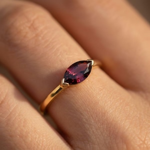 May include: A gold ring featuring a marquise-cut, deep red gemstone. The gemstone is set in a delicate gold band, creating a classic and elegant design. The ring is shown on a person's finger, highlighting its size and shape.
