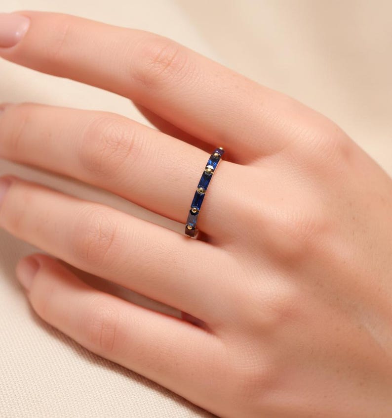 May include: A gold-toned ring with a band of alternating dark blue rectangular stones and small gold-colored dots. The ring is worn on a finger, showcasing its design and color contrast. The ring is a piece of jewelry.