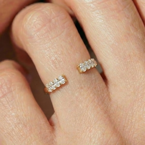 Puede incluir: Primer plano de dos anillos de oro, cada uno con una fila de pequeñas piedras preciosas transparentes. Los anillos son abiertos, con las filas de piedras preciosas a cada lado de un dedo. Las bandas de oro tienen un acabado pulido y las piedras brillan.