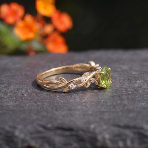 Könnte beinhalten: Ein goldener Ring mit einem zentralen, oval geschliffenen grünen Peridot-Edelstein. Die Ringschiene ist mit einem zarten Weinreben- und Blattmotiv gestaltet, das mit kleinen, klaren, rund geschliffenen weißen Steinen in den Zweigen verziert ist.