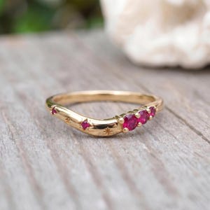 May include: A curved gold band ring featuring three round, vibrant red rubies set in a cluster on one side, complemented by smaller star-shaped engravings and a single tiny ruby accent embedded along the polished metal surface of the band.