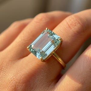 May include: A gold ring featuring a large, rectangular aquamarine gemstone. The light blue gemstone is set in a gold band with a simple, elegant design. The ring is shown on a finger.