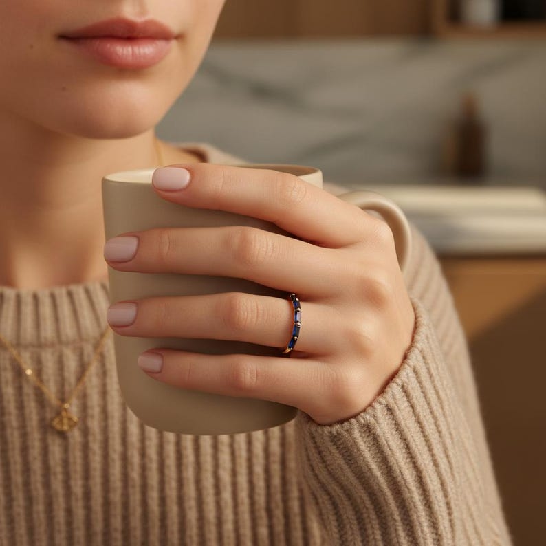 May include: A gold ring with a band of rectangular, dark blue gemstones. The ring is worn on a person's finger, and the person is holding a beige mug. The person is wearing a beige sweater and a gold necklace.
