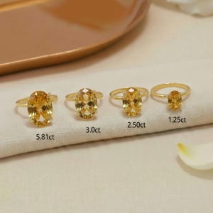 May include: Four gold rings with oval, yellow gemstones are displayed on a white cloth. The rings are labeled with their carat weights: 5.81ct, 3.0ct, 2.50ct, and 1.25ct. The background includes a gold tray and a white flower.