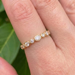 Op de afbeelding: Een gouden ring met een reeks in chaton gezette ronde en ovale heldere stenen, gerangschikt in een afwisselend patroon langs de bovenkant van de ring. Het metaal heeft een gepolijste geelgouden afwerking en de stenen zitten stevig in de zettingen.