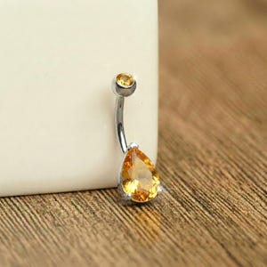 May include: A curved belly button ring with a silver-colored metal shaft. The ring features a small, round, yellow gemstone at the top and a larger, pear-shaped yellow gemstone at the bottom. The jewelry is set against a neutral background.