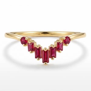 May include: A gold V-shaped ring featuring a series of seven vibrant red rubies. The design includes two round-cut rubies on the outer ends and five rectangular baguette-cut rubies arranged in a chevron pattern along the center of the band.