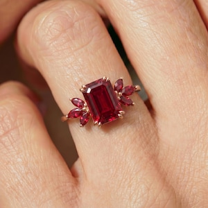 May include: A rose gold ring featuring a large emerald-cut deep red gemstone as the center stone, flanked on each side by three smaller marquise-cut red gemstones arranged in a cluster, set on a thin, polished rose gold metal band.