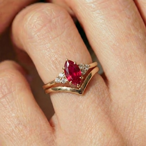 May include: A gold ring set featuring a marquise-cut ruby gemstone and small accent diamonds. The ring is paired with a chevron-shaped band, creating a layered look. The ruby is a vibrant red color.