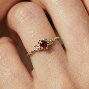 May include: A gold ring featuring a floral design with a central red gemstone. The ring has delicate leaf accents and small, sparkling stones along the band, creating an elegant and nature-inspired aesthetic.