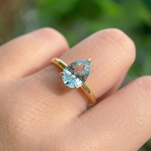 以下が含まれることがあります： 洋梨型の水色の宝石をあしらったゴールドリング。 宝石は4つの爪で固定されています。 リングのバンドはシンプルなポリッシュゴールドです。 クラシックなデザインです。